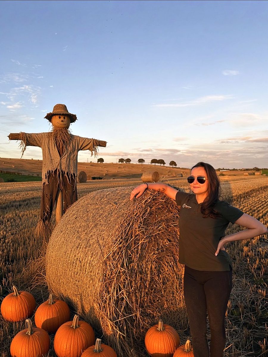 Katie Lunn, registered veterinary nurse, leaning on a hay bale beside a friendly scarecrow and pumpkins at sunset